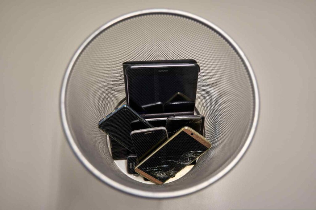 Several broken and discarded smartphones, including one with a shattered screen, piled inside a metal mesh trash bin.