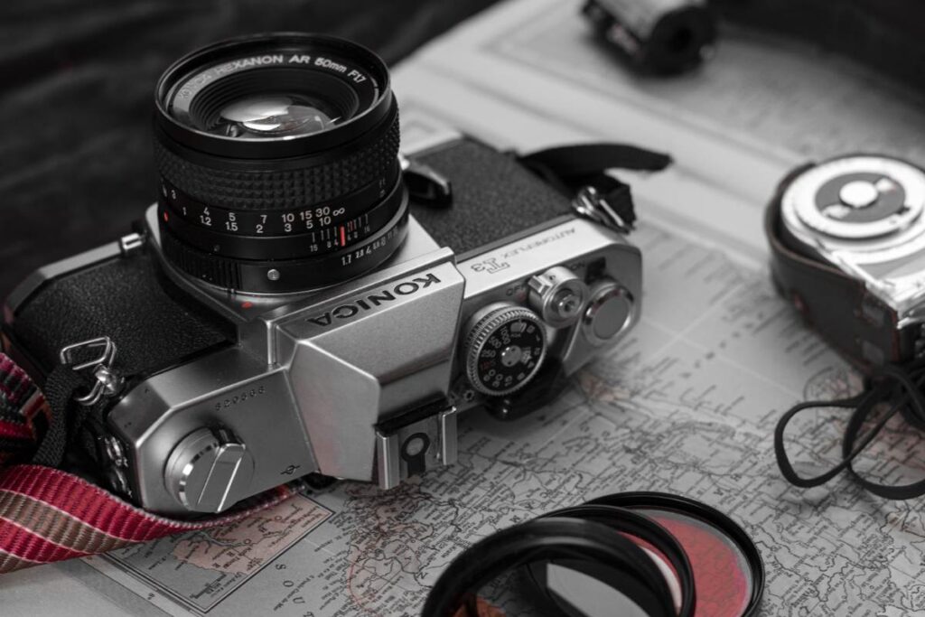 Silver Konica Autoreflex T3 film camera with a 50mm lens, resting on a paper map next to a camera strap and lens filter.